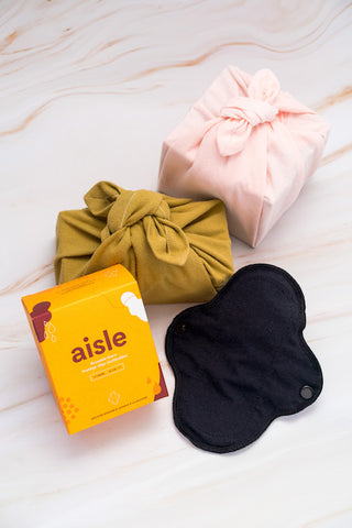 A black reusable liner displayed beside an orange box labelled Aisle Reusable Liners, and two other small decorative boxes wrapped in yellow and pink fabric.