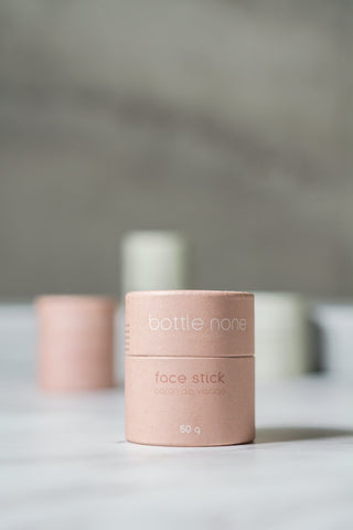 A pink paper-based tube labelled as a Face Stick from the brand Bottle None. The stick is displayed on a white surface, with 3 other pink and green tubes blurred in the background.