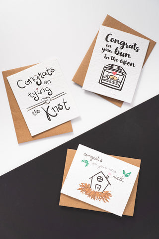 Three congratulatory greeting cards with their respective kraft envelopes laid out on a black and white background. 