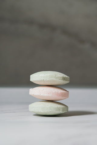A stack of three face wash bars in alternating colours of mint green and pink, on a white surface in front of a concrete background. 