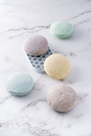 An assorted arrangement of shampoo bars in mint green, lavender, yellow, blue and grey, arranged on a marble surface.