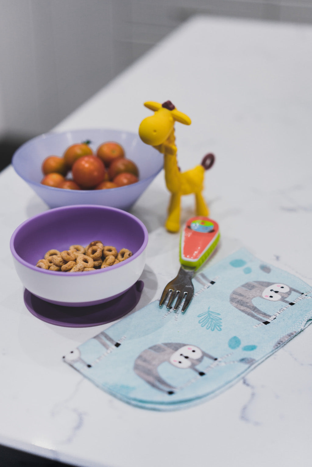 Cheerios and cherry tomatoes in children's bowls with fork, toy giraffe and printed cotton napkin