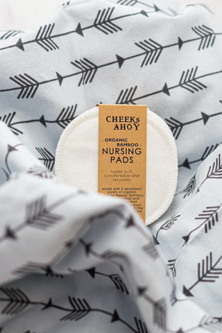 A pack of white organic bamboo nursing pads wrapped in minimal kraft paper band, nestled in a grey fabric with arrow patterns.
