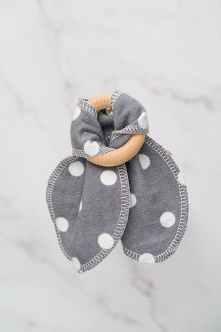 White polka dots on grey cotton teething ears that are attached to a maple wood ring, in front of a marble background.