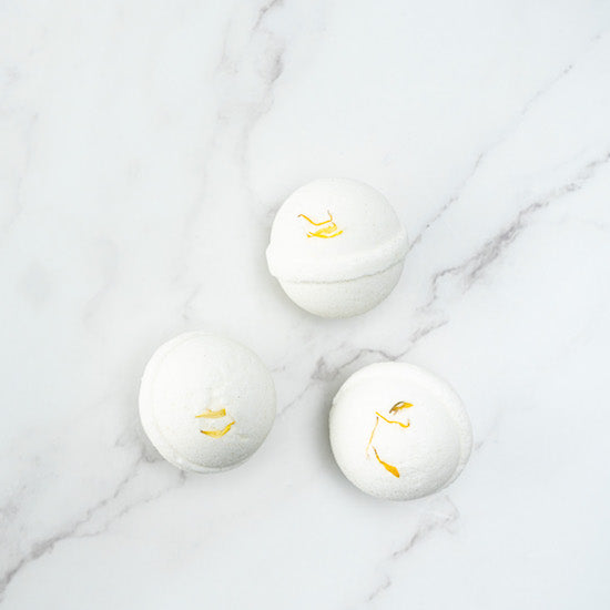 Three white coloured bath bombs with yellow dried calendula petals on top, displayed on a marble surface.