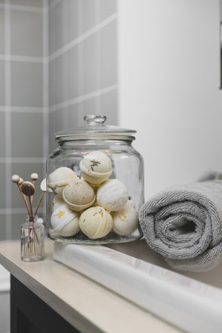 A large glass jar filled with bath bombs sitting on the edge of the sink in the bathroom, beside a rolled up grey towel and decorative accessories on the sink.