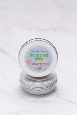 Two circular tin containers labelled Baby Butt Balm by Old Soul Soap Company, displayed on a marble surface.