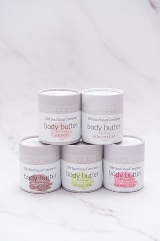 Five grey cylindrical paper tube containers each labelled Body Butter in various scents by Old Soul Soap Company, stacked and displayed on a marble surface.