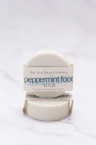 Two round white bars, each with minimal paper packaging labelled Peppermint Foot Scrub by Old Soul Soap Company, stacked and displayed on a marble surface.