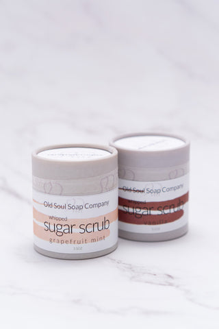Two cylindrical paper-tube light grey containers labelled as Whipped Sugar Scrub, one in Grapefruit Mint and the other in Vanilla scent, by company Old Soul Soap Company, displayed on a marble surface.