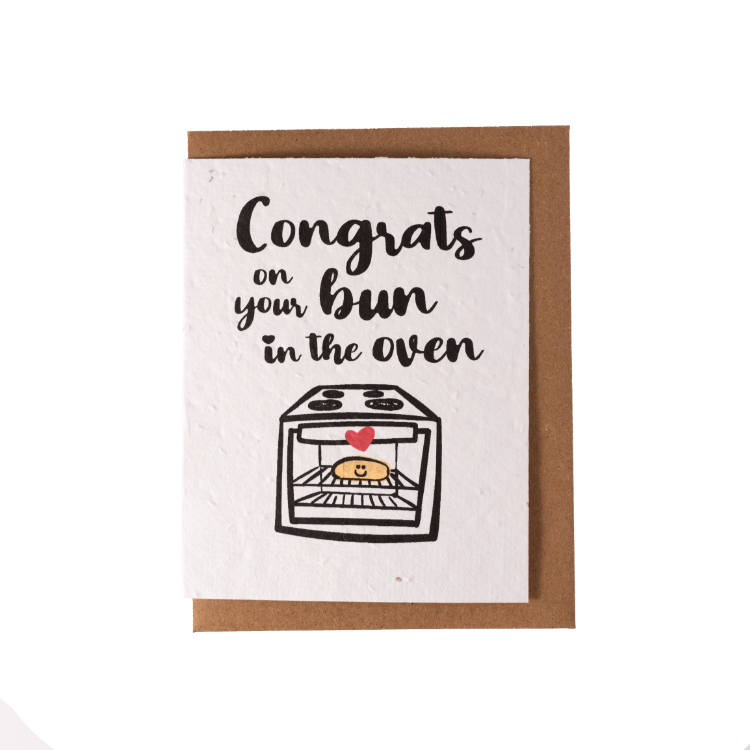 A white greeting card laying over top of a kraft envelope. The card contains an image of a small oven with a bun inside, with text "Congrats on your Bun in the Oven"