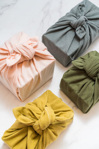 Four boxes of various sizes each wrapped in a different coloured Furoshiki fabric gift wrap in nude pink, light heather grey, olive grass and lemongrass colour. 