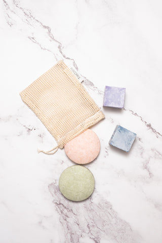 A mesh cotton soap bag with four various coloured bars of soap placed just outside its drawstring opening and around it.